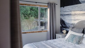 Une chambre avec une grande fenêtre, un lit avec des oreillers, une table de chevet avec une lampe et un tableau représentant un paysage marin.