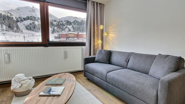 Ein modernes Wohnzimmer mit grauem Sofa, einem Holztisch und Blick auf eine verschneite Berglandschaft durch große Fenster.