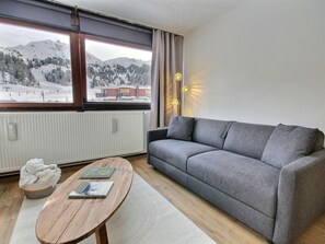 A modern living room with a grey sofa, a wooden coffee table, and a view of a snowy mountain landscape through large windows. - Renovated studio cabin at the foot of the slopes (La Plagne)