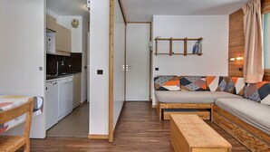 Ett kompakt bostadsutrymme med ett litet kök, en soffa med mönstrade kuddar, en träbänk och en vägghylla.