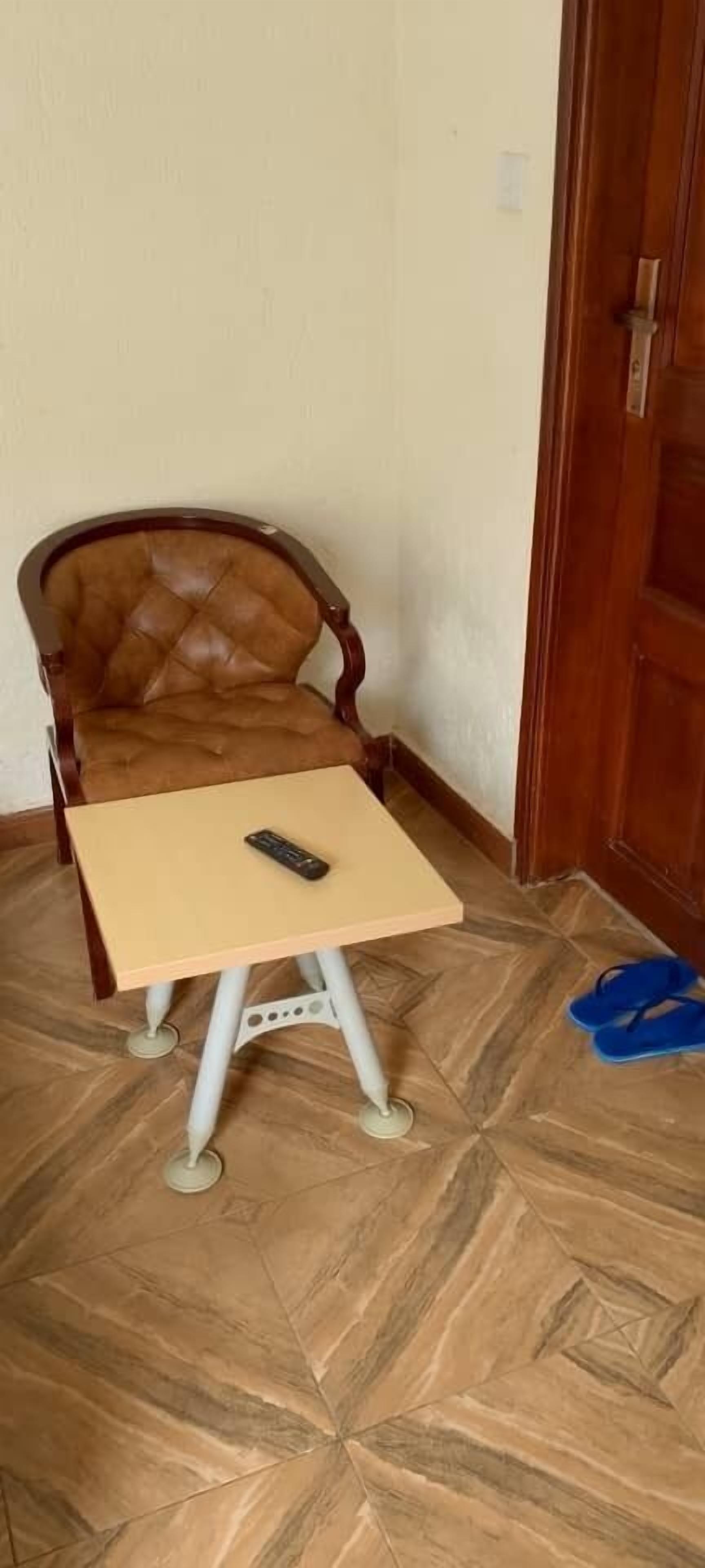 Una stanza con una sedia di legno, un tavolino con un telecomando e un paio di pantofole blu sul pavimento.