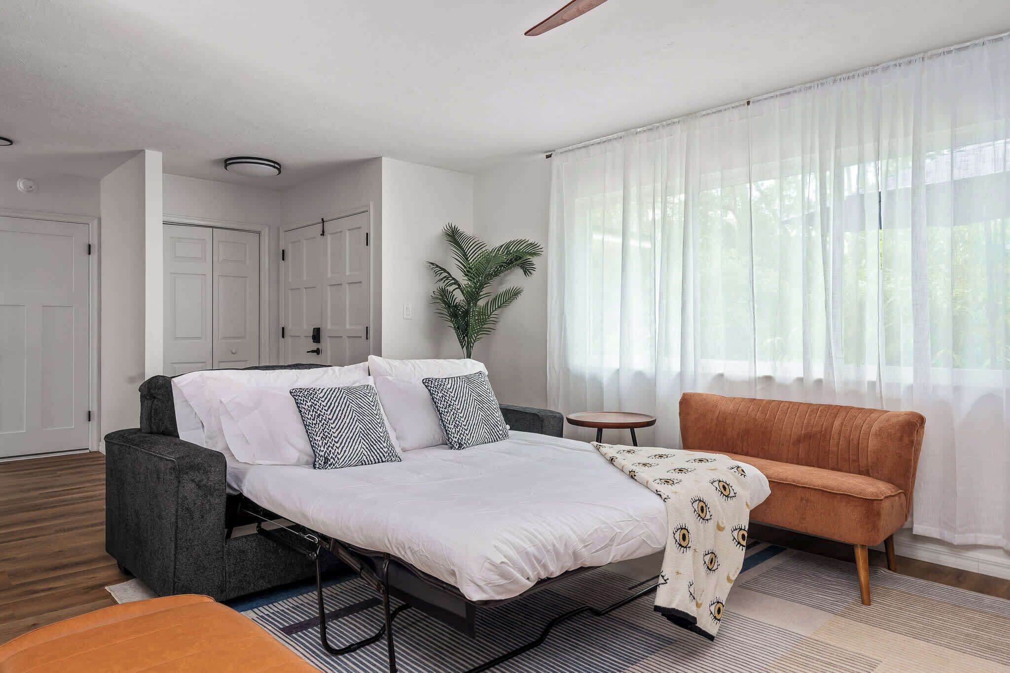A modern hotel room with a bed, a grey sofa, a small round table, and a large window with sheer curtains.