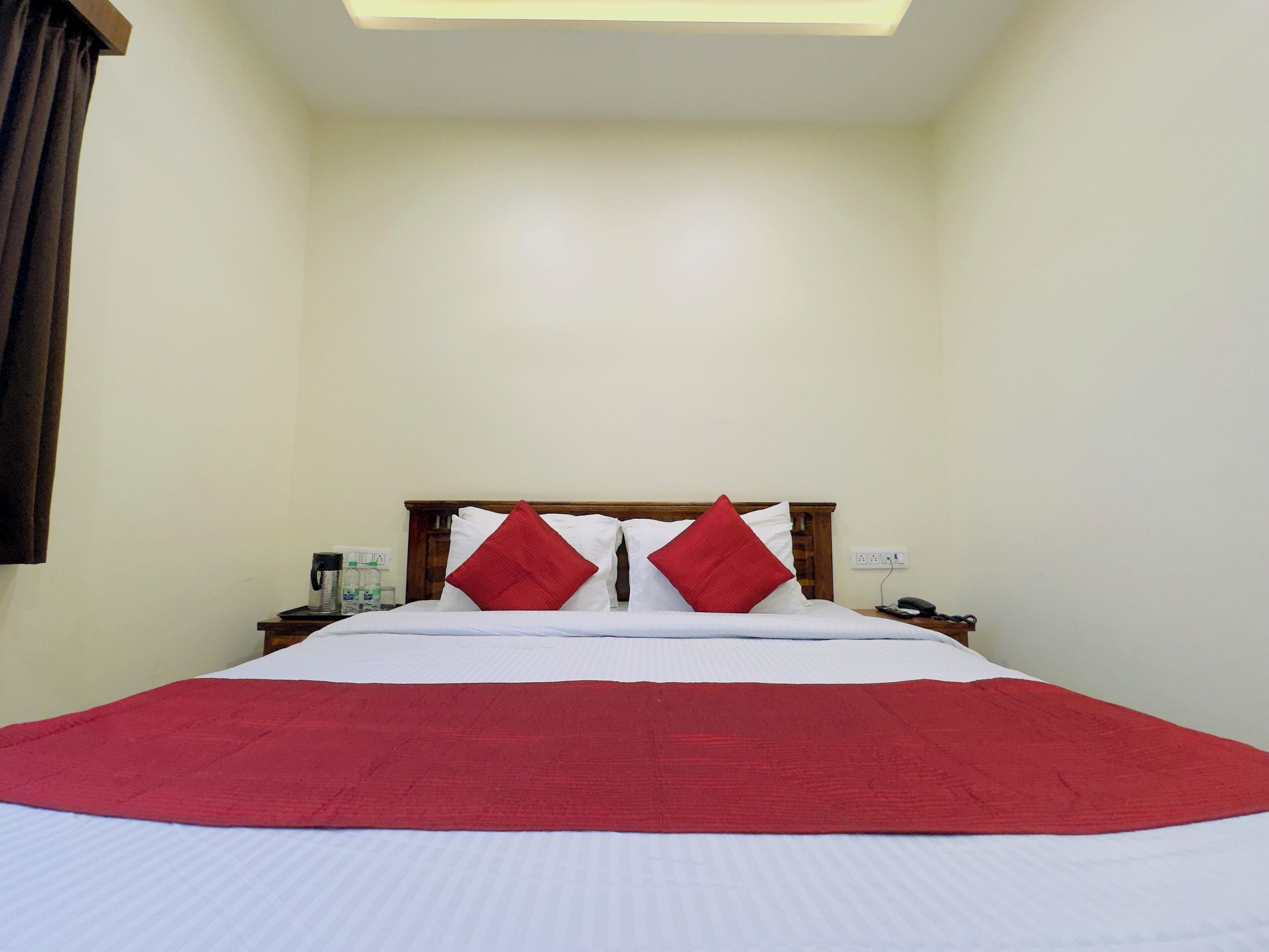 A neatly made bed with white and red bedding, a wooden headboard, and a bedside table with a lamp and phone.