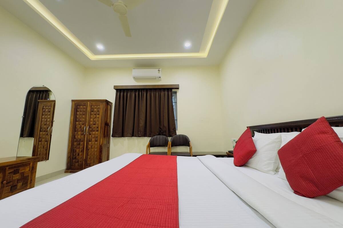 Habitación de hotel con una cama grande, ventilador de techo, equipo de aire acondicionado, armario de madera, una silla y una ventana con cortinas.