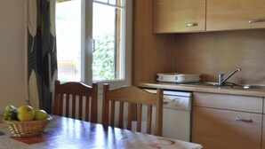 A kitchen with wooden cabinets, a dining table with chairs, a basket of fruit, and a window with curtains. - Chalet 1 en Cévennes (Aulas)