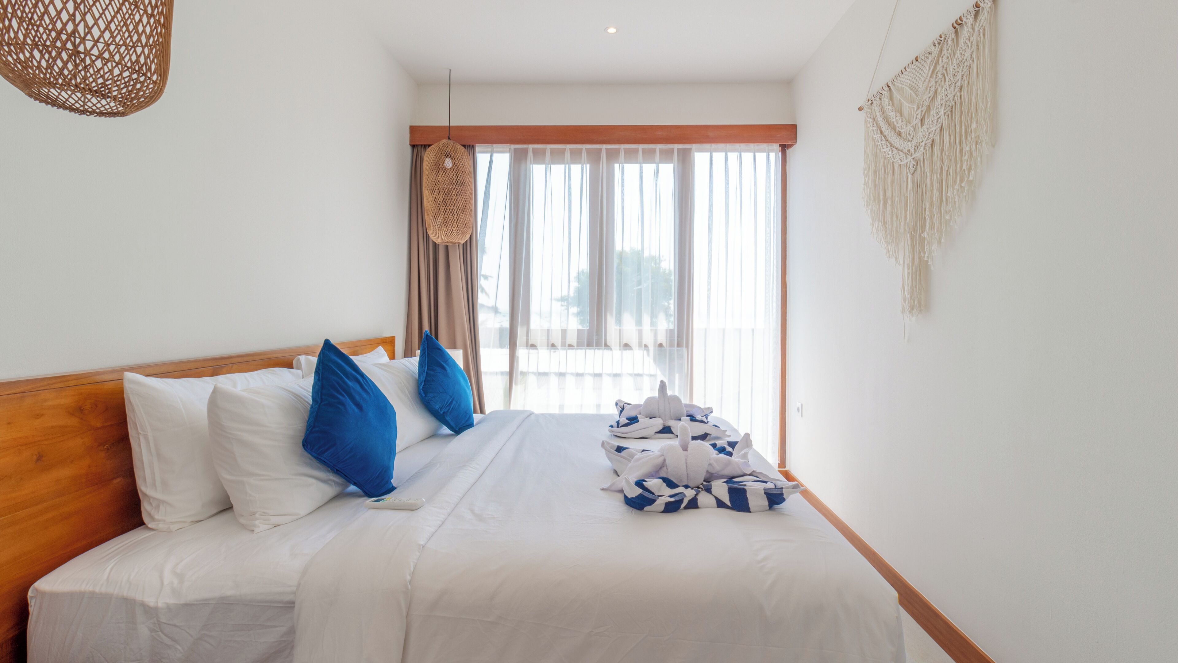 A hotel room with a bed, white bedding, a wooden headboard, and a view of the outdoors through large windows.
