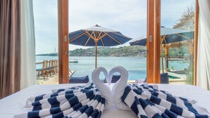 A bed with a blue and white checkered blanket and two swan-shaped pillows. Outside, there are two umbrellas and a view of a body of water with boats. - Segara Seaside Resort (Ceningan Island)