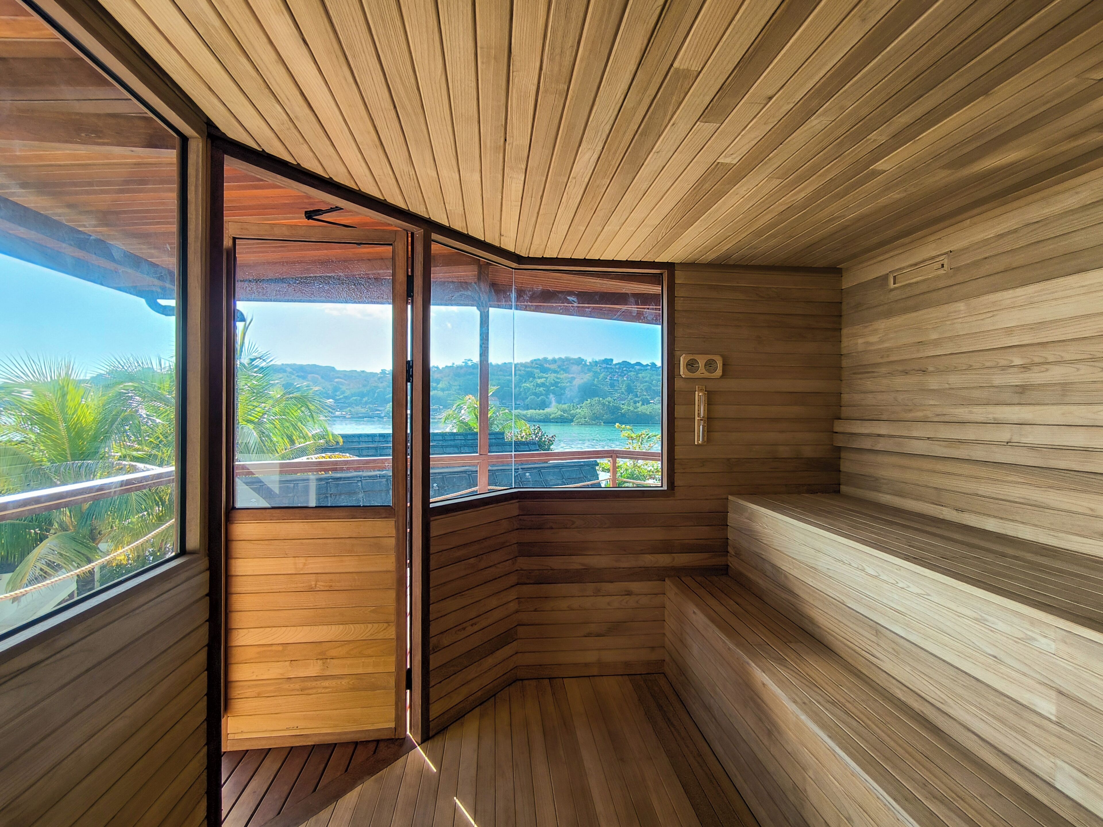 A sauna room with wooden walls and a large window offering a view of a tropical landscape.
