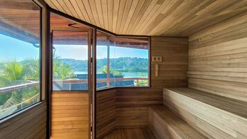 A sauna room with wooden walls and a large window offering a view of a tropical landscape.