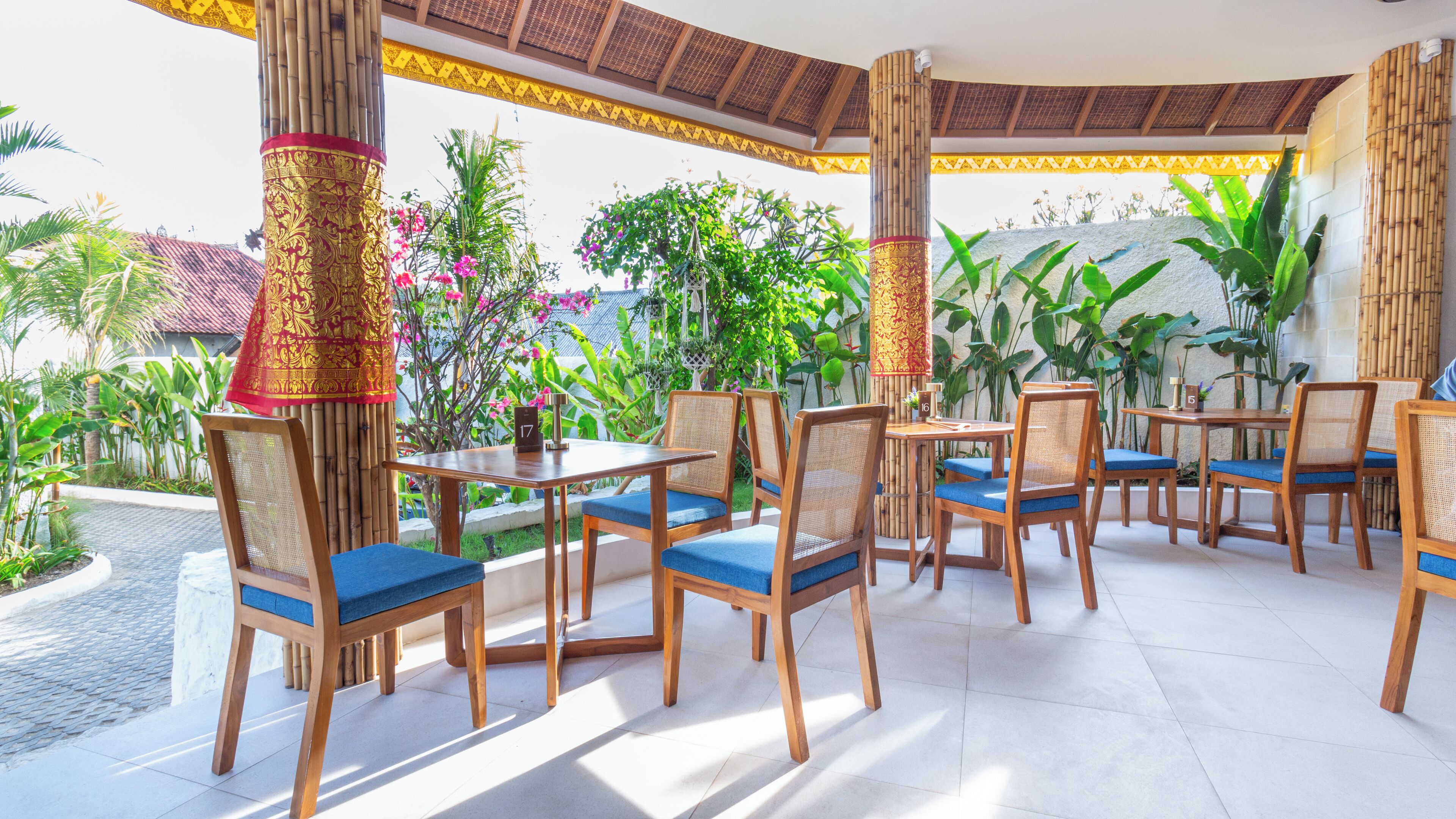 An outdoor dining area with wooden tables and chairs, surrounded by tropical plants and a decorative bamboo structure.