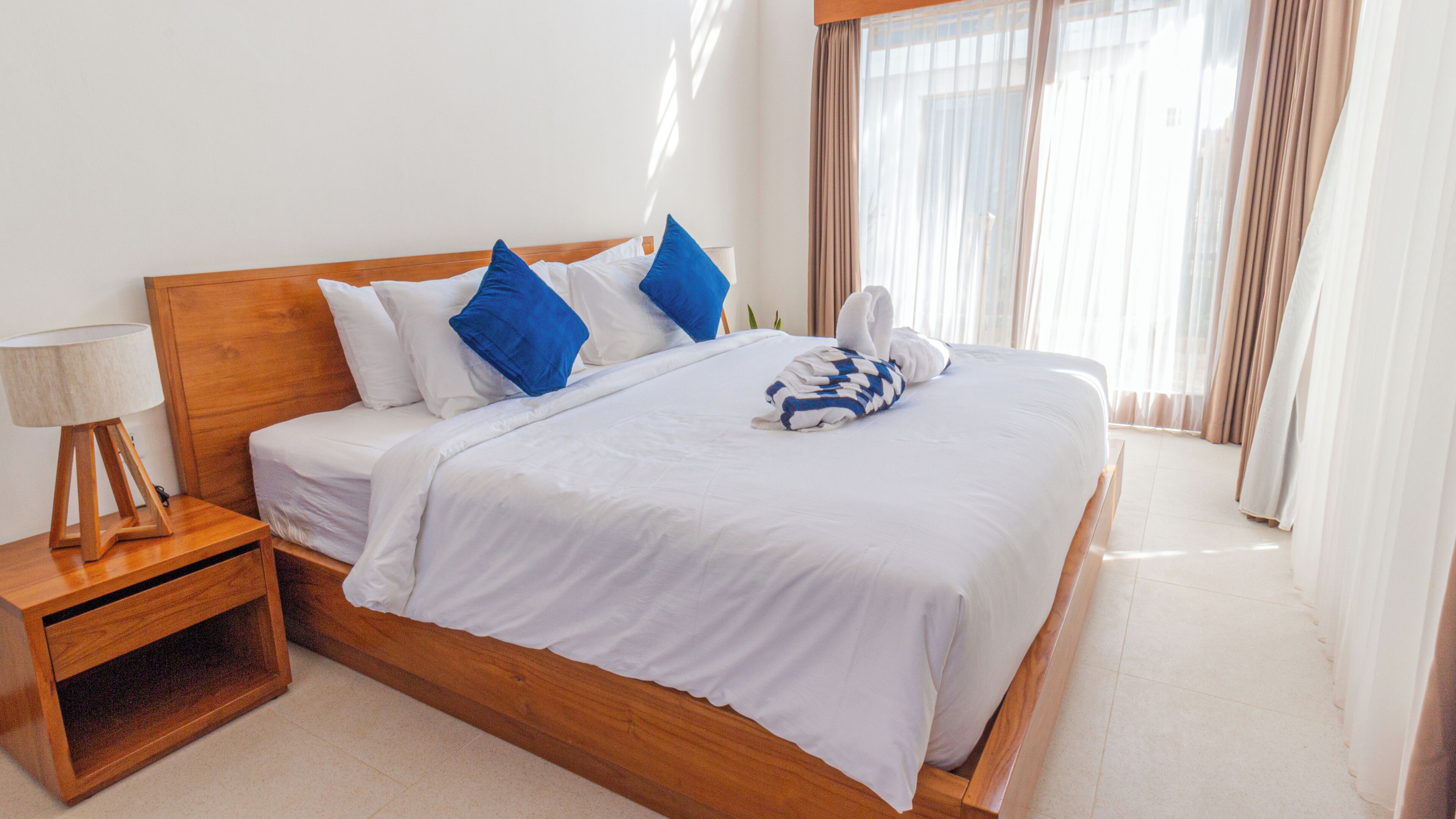 Uma cama bem arrumada com roupa de cama branca e almofadas azuis, uma mesa de cabeceira de madeira com uma luminária e uma janela com cortinas finas.