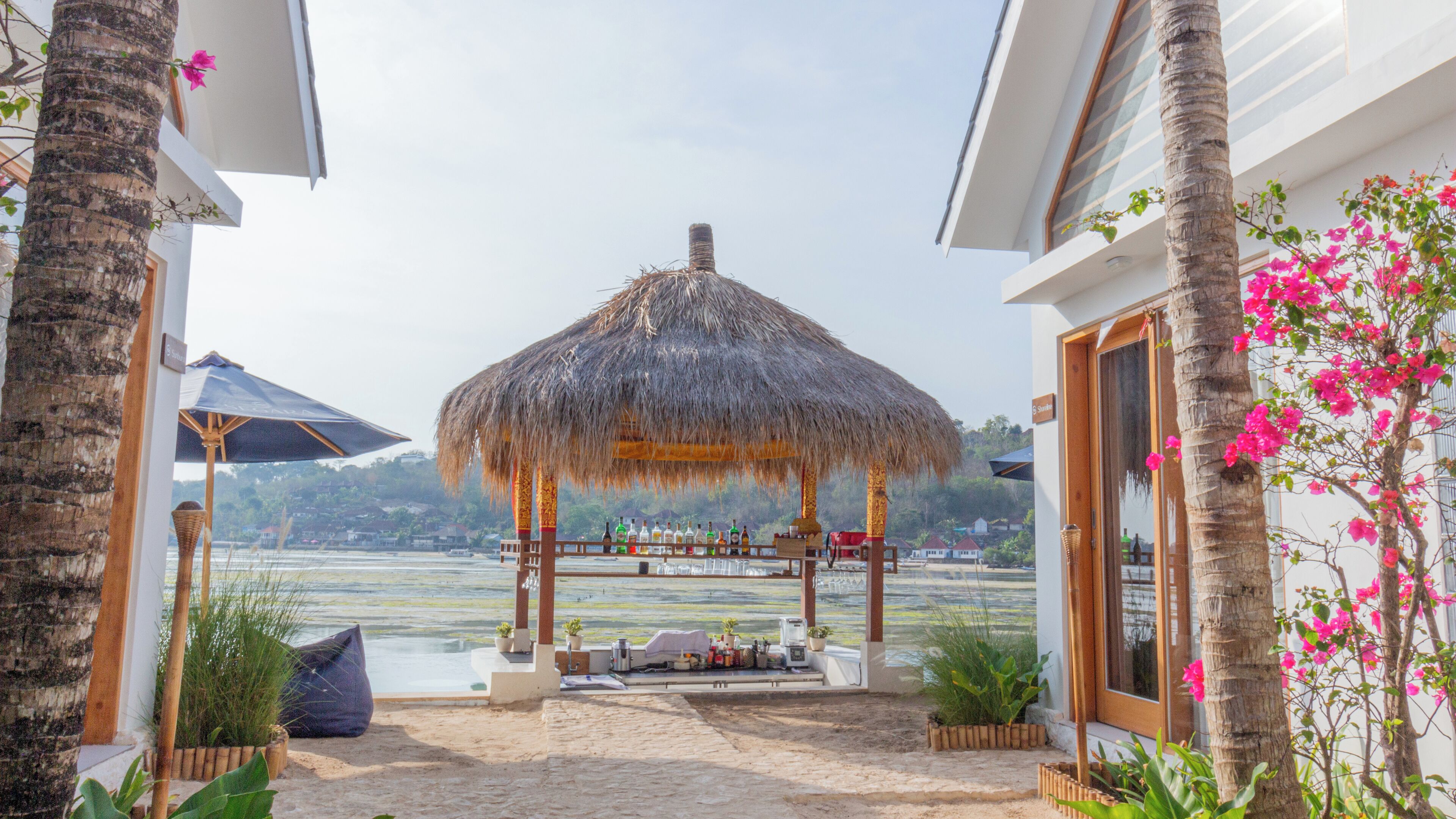 A thatched gazebo with a table set for outdoor dining, overlooking a beach and water.