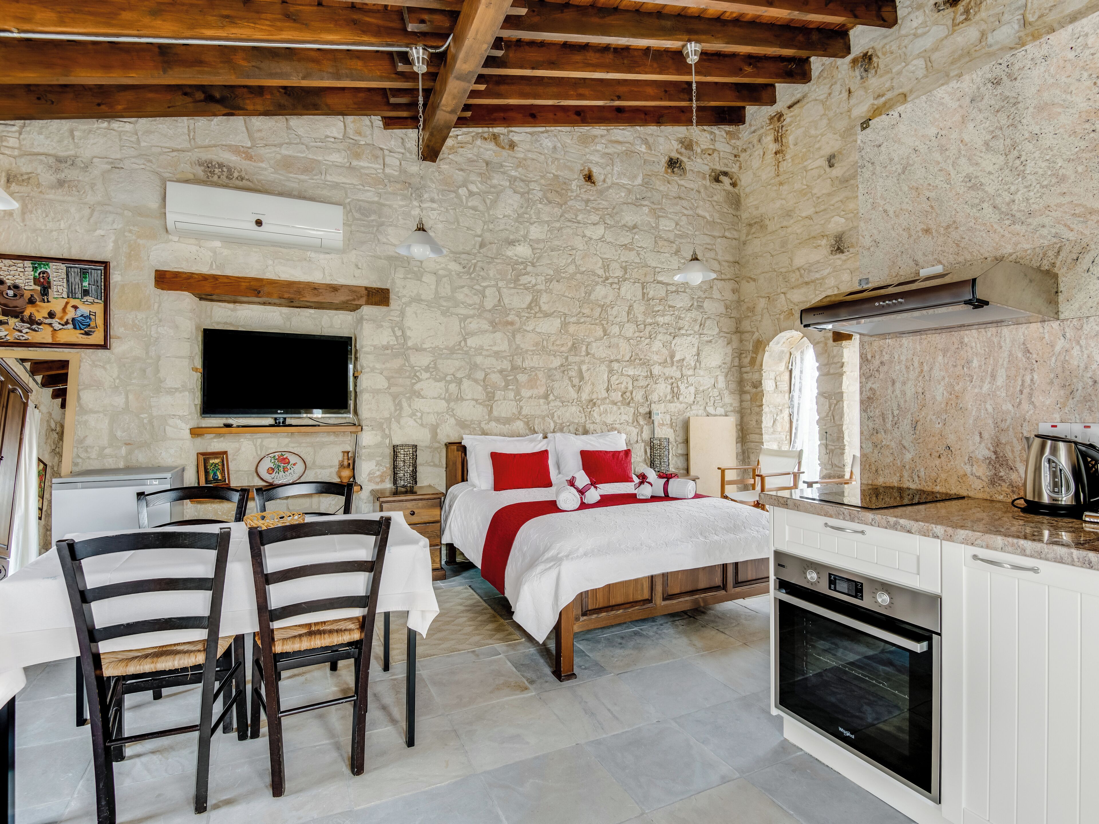 A cozy room with a bed, a TV, a dining table with chairs, a kitchen area with an oven, and stone walls.
