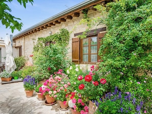 A stone building with a wooden balcony and a window with shutters. There are potted plants with flowers in front of the building. - Arsorama Village Homes (Arsos)