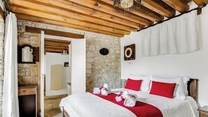 A bedroom with a stone wall, wooden ceiling beams, a bed with white and red bedding, a wooden nightstand, and a stone fireplace. - Arsorama Village Homes (Arsos)