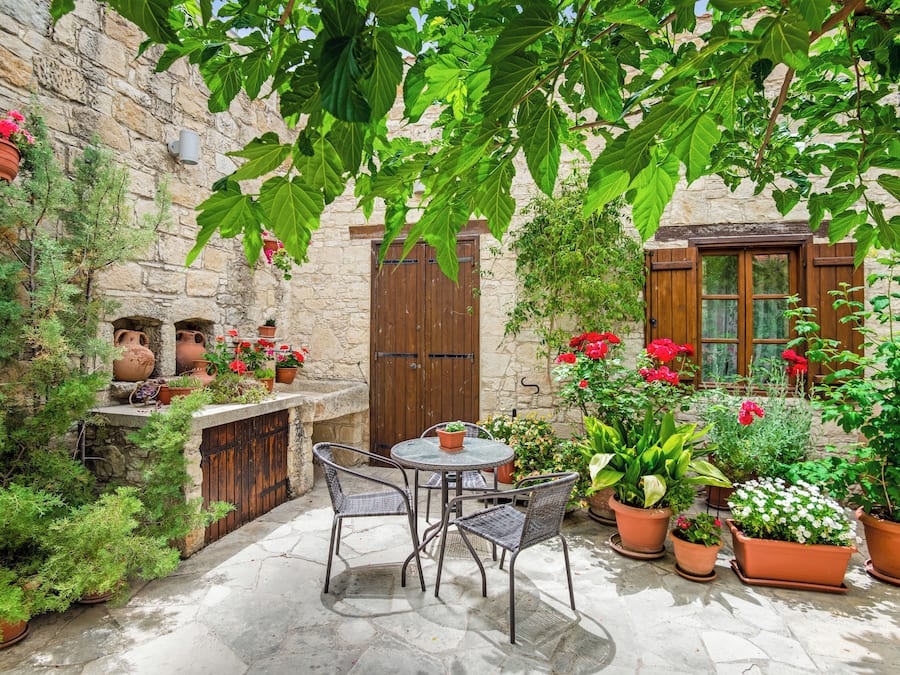 Un pati de pedra amb una taula i cadires, envoltat de plantes i flors en testos.