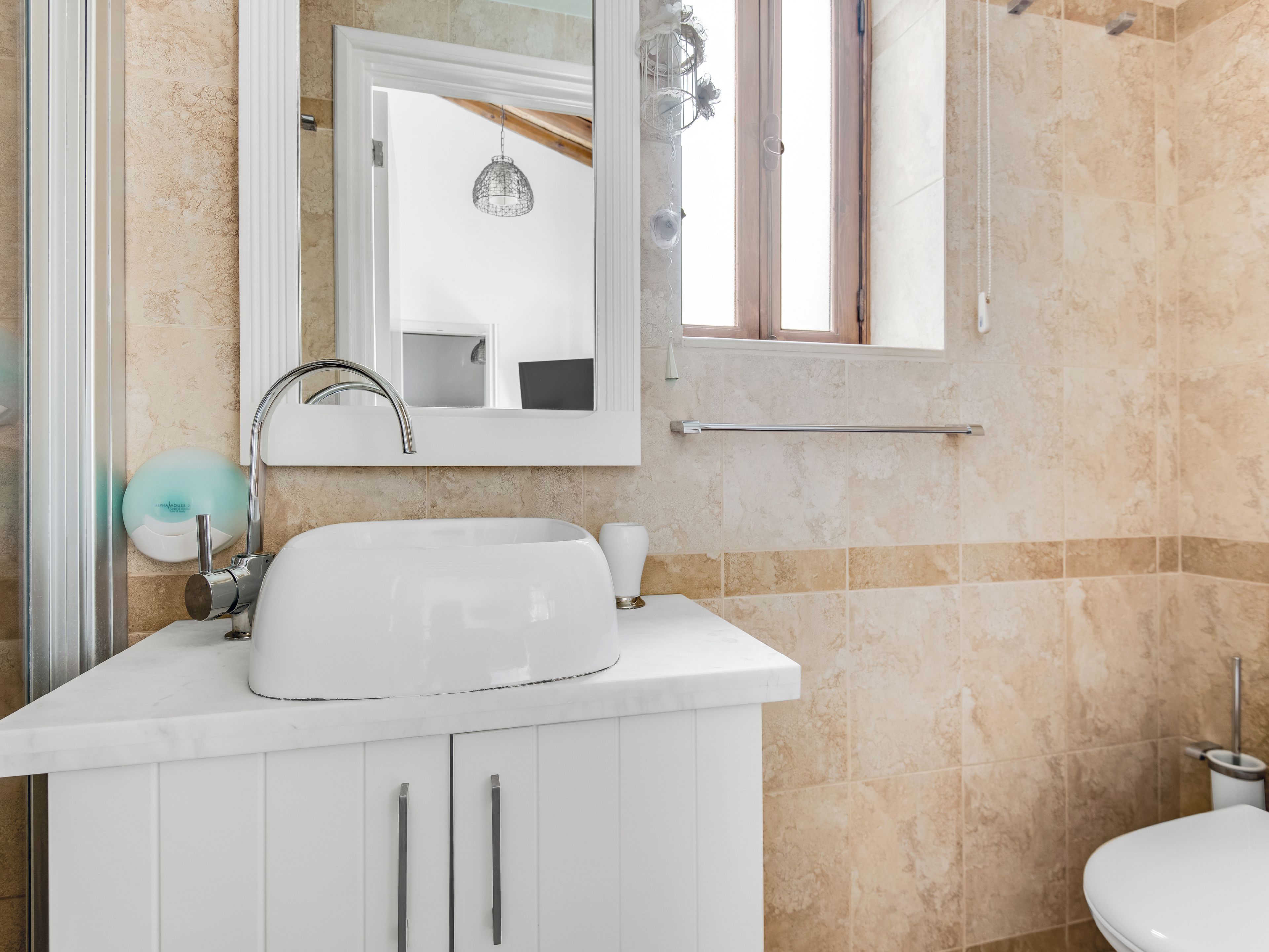 Un baño con lavamanos blanco, espejo y mueble. Hay un perchero sobre el lavamanos y un inodoro a la derecha.