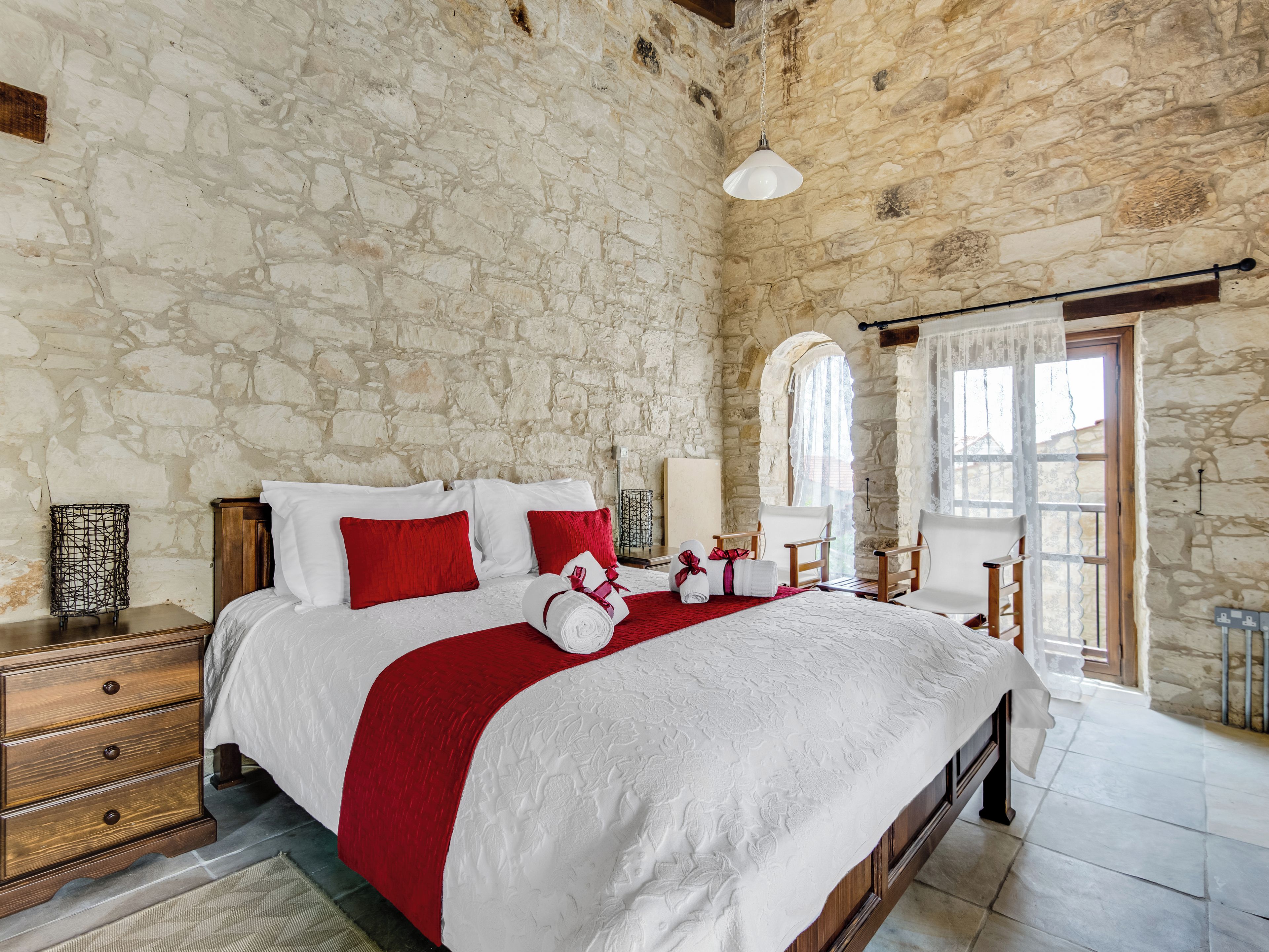 A stone-walled bedroom with a large bed, red pillows, and a wooden dresser.