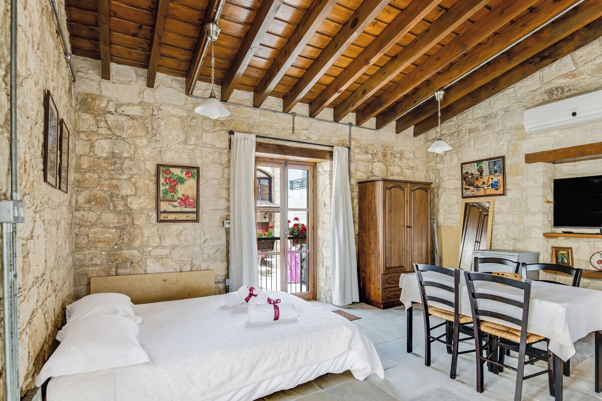 A room with a bed, wooden ceiling beams, stone walls, a dining table with chairs, and a television.