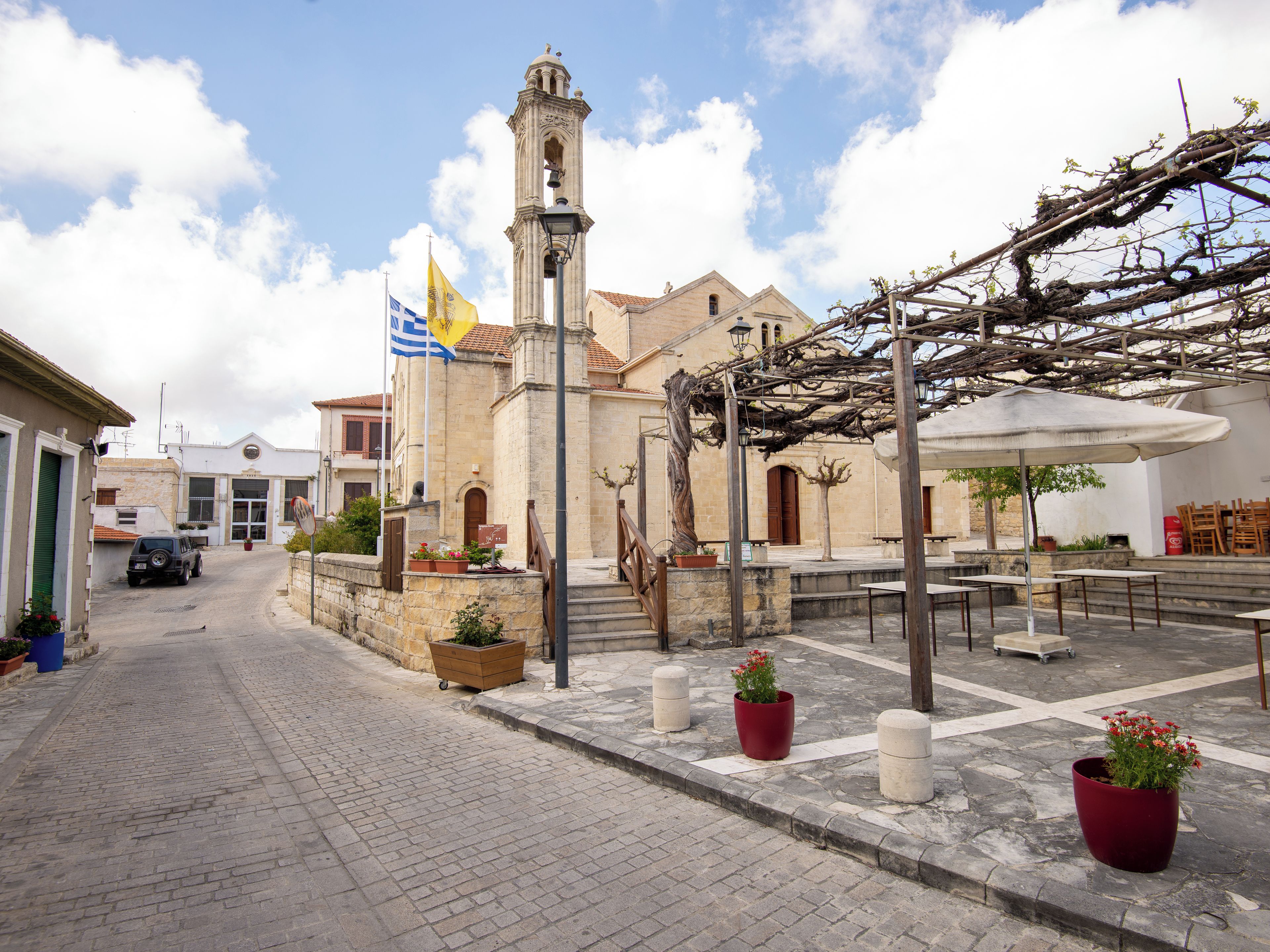 Uma rua de paralelepípedos com um edifício histórico, torre sineira, mesas ao ar livre e plantas em vasos.