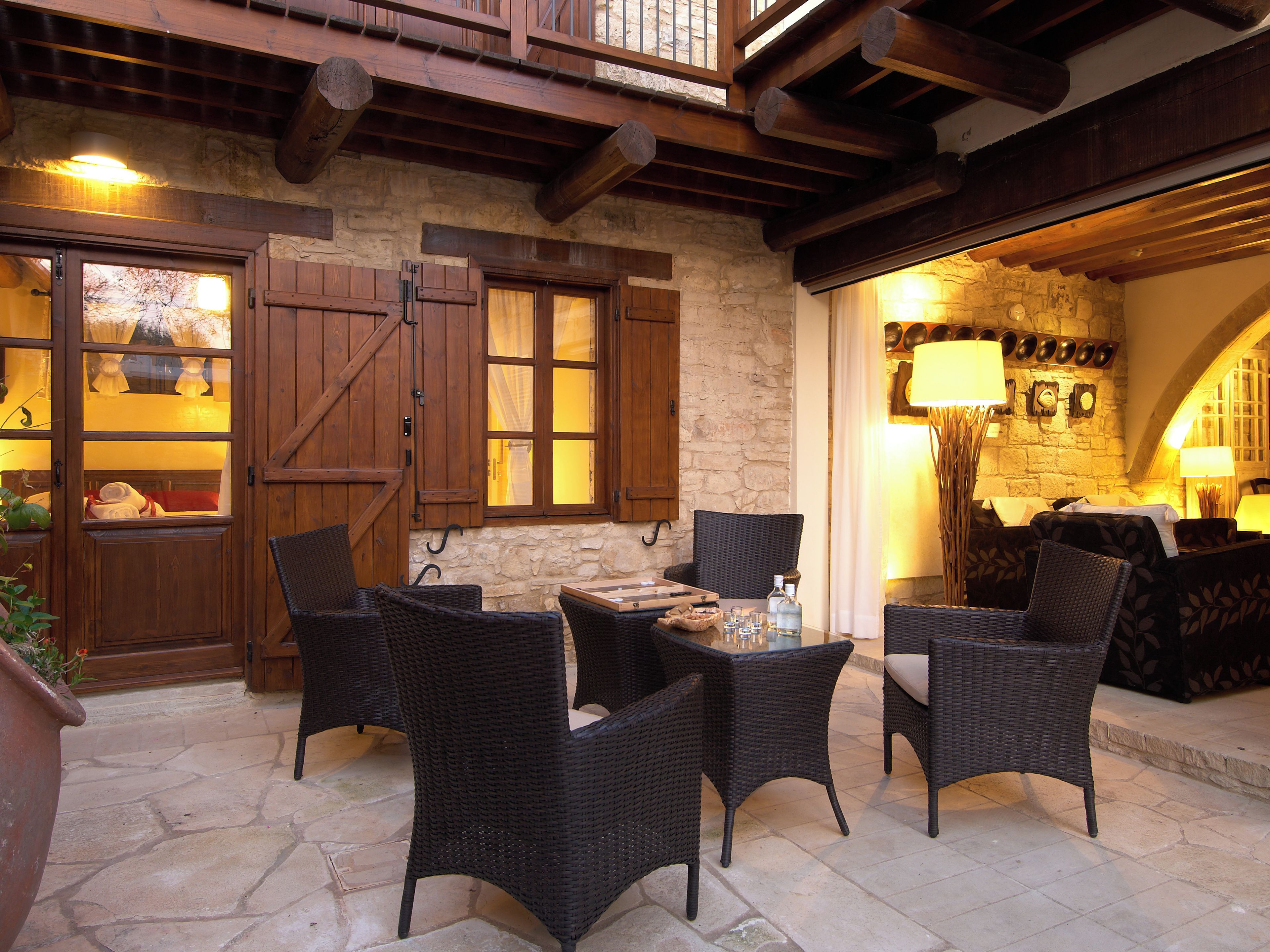 Un patio con paredes de piedra, muebles de mimbre, una mesa con vasos y una botella, y contraventanas de madera en la puerta.