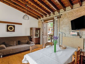 A cozy living room with a wooden ceiling, a stone wall, a wooden cabinet, a sofa, a dining table with a white tablecloth, and a green vase. - Arsorama Village Homes (Arsos)
