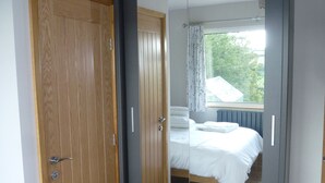 Un dormitorio con armario de madera, puerta de armario con espejo, cama con ropa de cama blanca y ventana con cortinas.