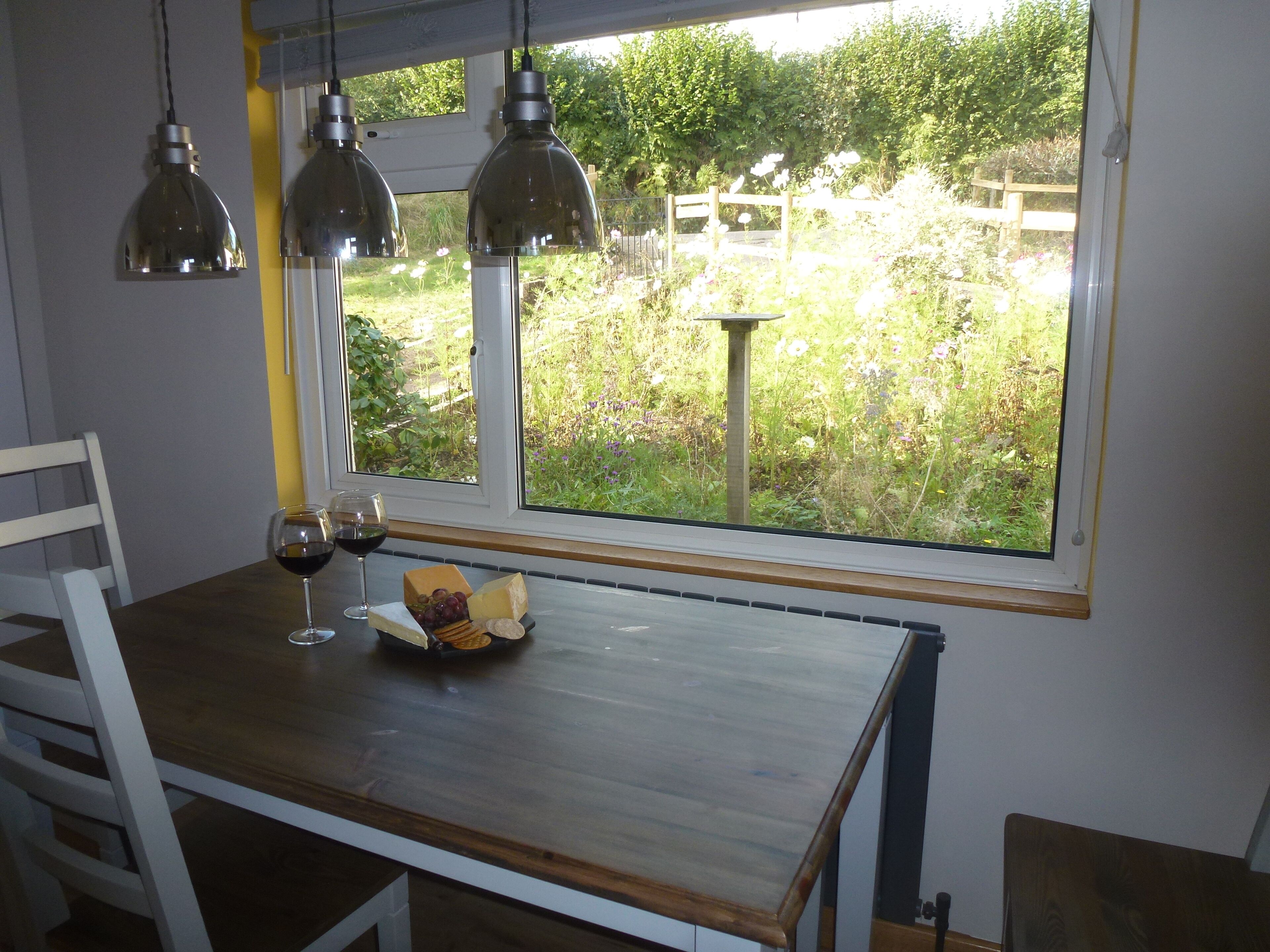 Une table à manger avec un verre de vin et du fromage, une fenêtre donnant sur un paysage verdoyant et des lampes suspendues.