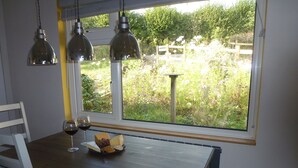 Una mesa de comedor con una copa de vino y queso, una ventana con vista a follaje, y lámparas colgantes.