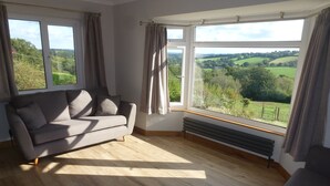 Una sala de estar moderna con un sofá gris, un radiador y ventanas grandes que ofrecen vistas a un paisaje verde.