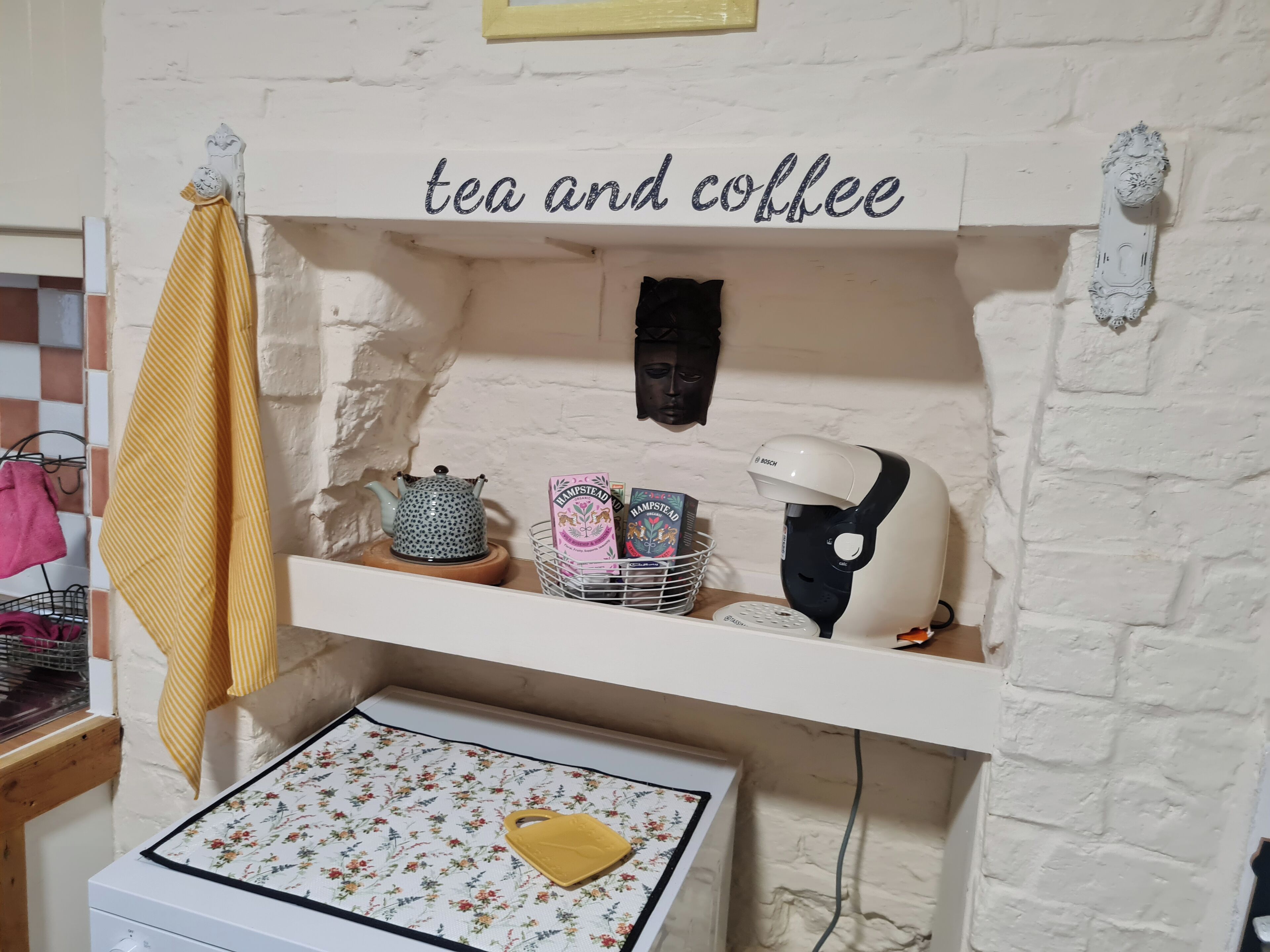 A kitchen area with a shelf labeled "tea and coffee," containing a teapot, coffee maker, and tea bags.