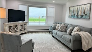 Un salón con un sofá gris, un sillón blanco, un televisor de pantalla plana sobre un mueble de madera y un cuadro de veleros en la pared.