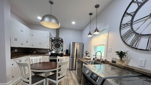 Una cocina moderna con armarios blancos, una mesa de comedor con sillas, un reloj de pared grande, electrodomésticos de acero inoxidable e iluminación colgante.