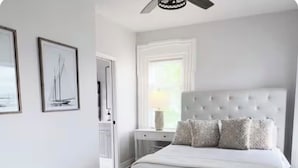 Un dormitorio con cama, mesita de noche, ventana y cuadros en la pared.