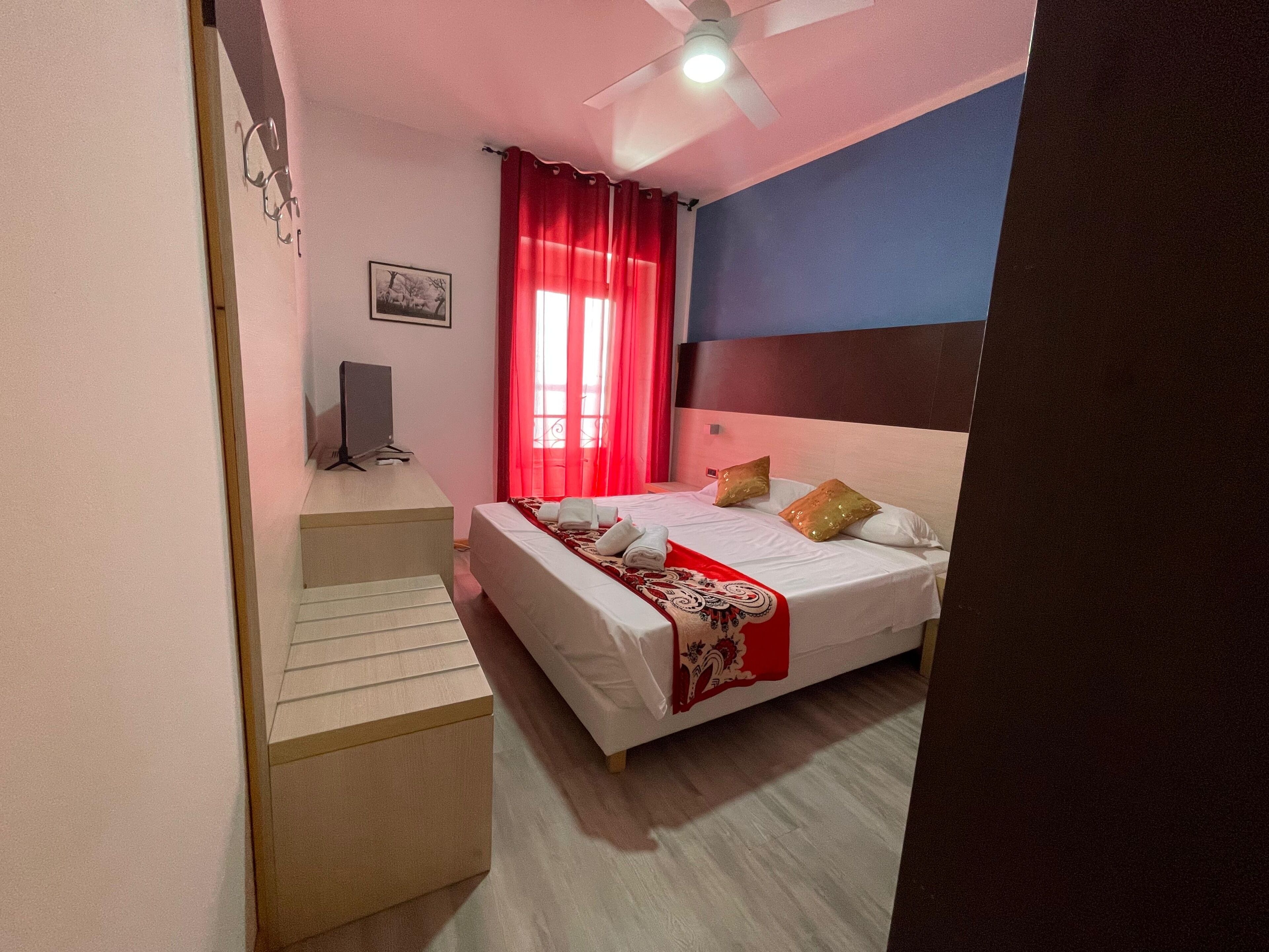 Quarto com cama, escrivaninha, cadeira, ventilador de teto e janela com cortinas vermelhas.