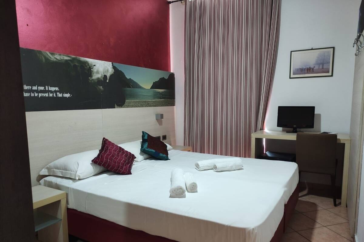 A hotel room with a bed, pillows, a desk with a computer, a chair, and a curtain.