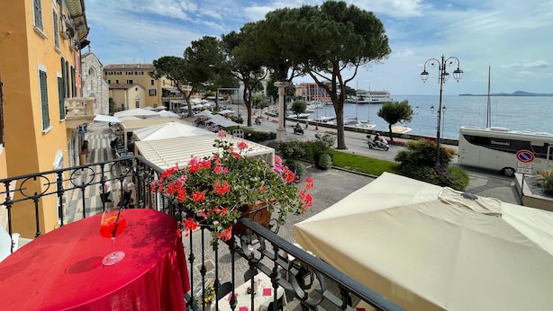 Un balcón con mantel rojo, un arreglo floral y vista a un puerto deportivo con barcos y un edificio al fondo.
