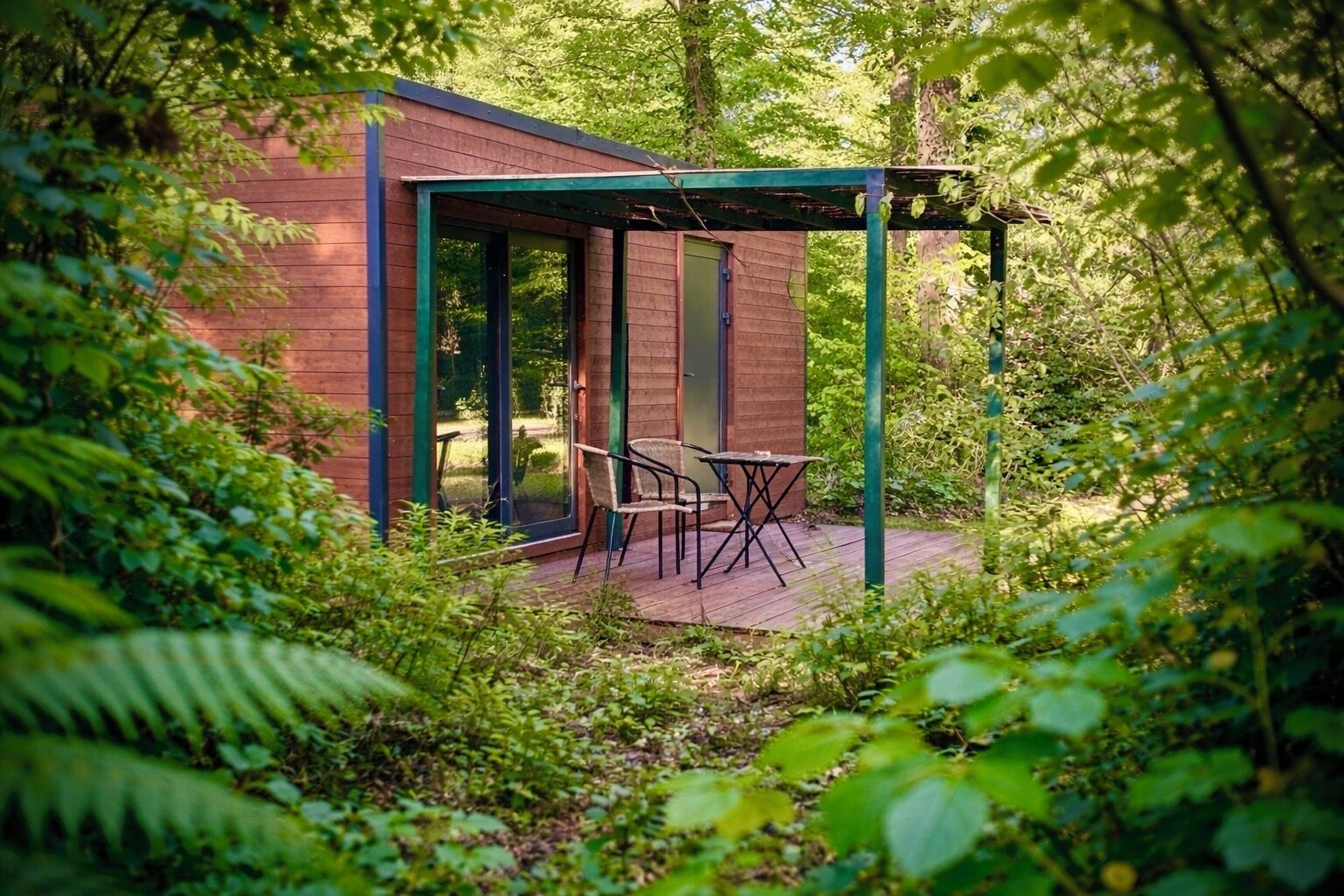 A wooden cabin with a glass door, a small patio with a table and chairs, surrounded by dense greenery.