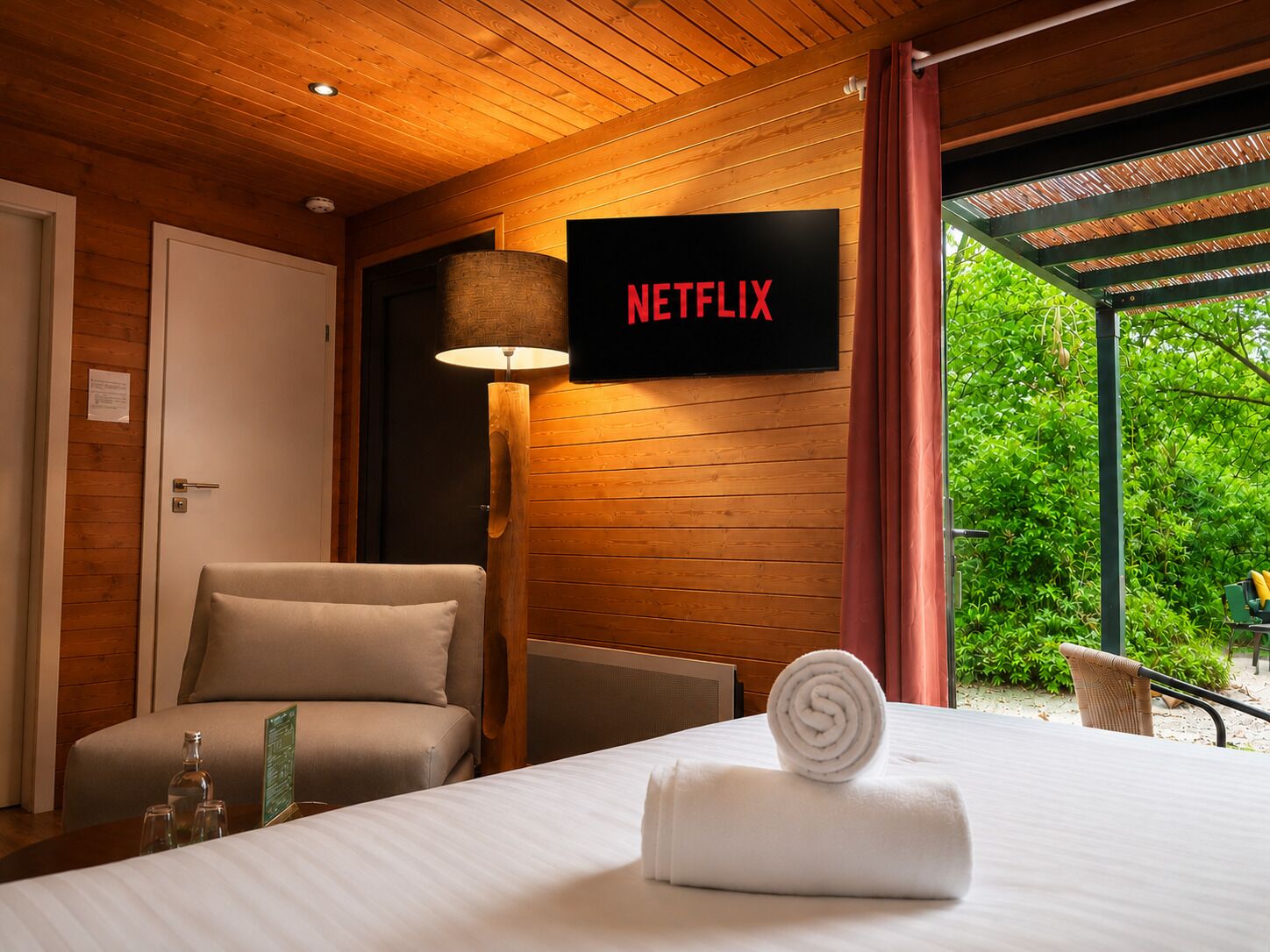 A hotel room with a bed, a chair, a lamp, a sign for Netflix, and a view of greenery.