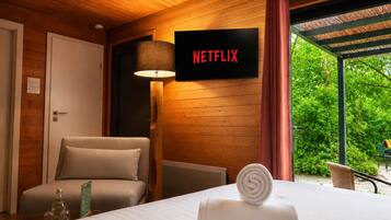 A hotel room with a bed, a chair, a lamp, a sign for Netflix, and a view of greenery.