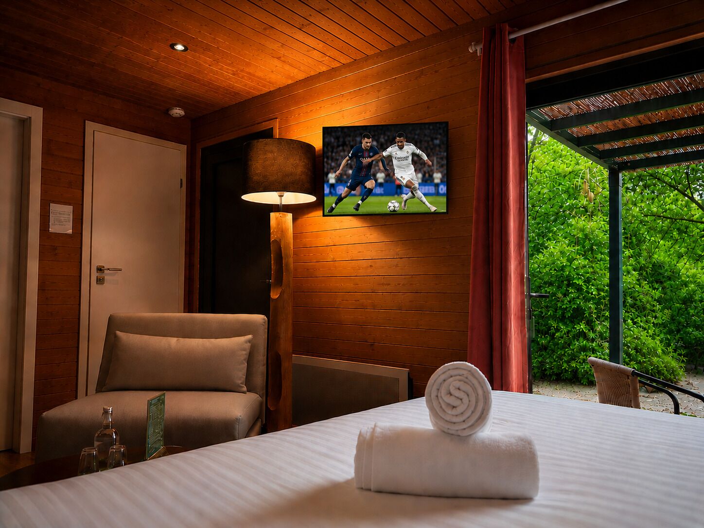 A hotel room with a bed, a television mounted on the wall, a chair, a lamp, and a view of greenery outside.