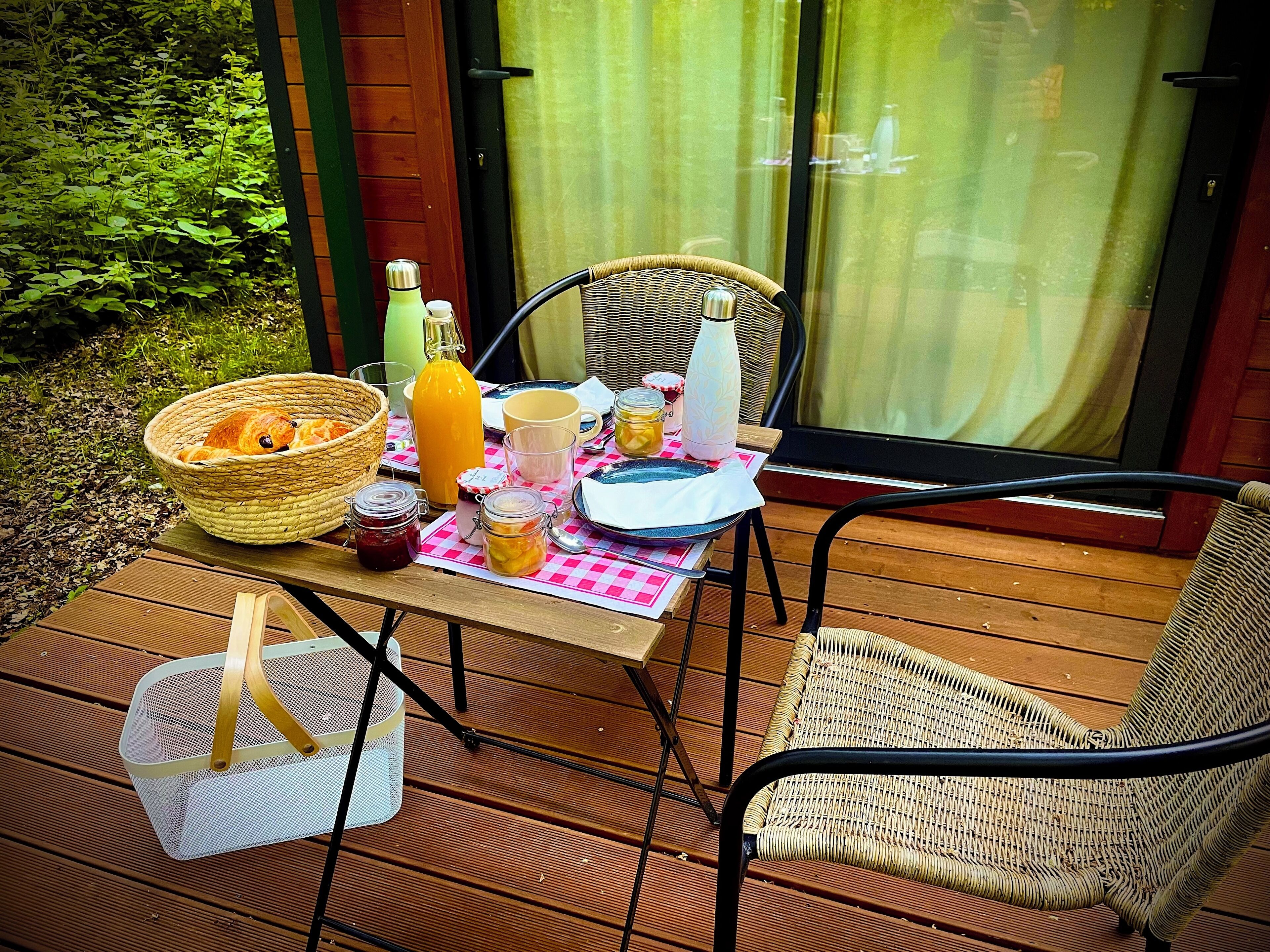En udendørs spiseplads med fletmøbler, et bord dækket med mad og drikke, og en kurv med en kat.
