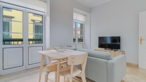 Ett modernt vardagsrum med en grå soffa, ett matbord i trä med stolar, en tv på ett träställ och stora fönster med persienner.