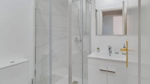 Ett modernt badrum med en dusch som är avskärmad med glasväggar, en vit tvättställ med ett guldfärgat blandare och en spegel ovanför tvättstället.