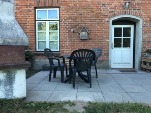 A brick building with a white door and windows, a small outdoor seating area with plastic chairs and a table, and a brick wall with a decorative birdhouse. - Das Abendrote Haus (Suesel)