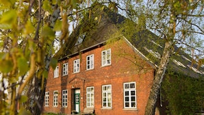 A red brick building with multiple windows and a green door, surrounded by trees and a grassy lawn. - Das Abendrote Haus (Suesel)
