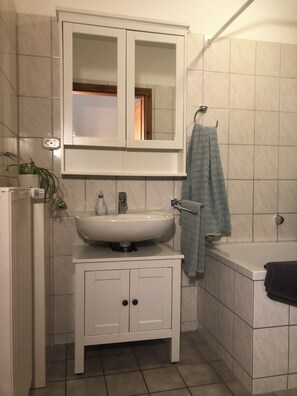 A bathroom with a white sink, mirror, and cabinet, a towel rack, and a bathtub. - Das Abendrote Haus (Suesel)
