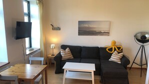 A living room with a dark-colored sofa, a white coffee table, a wooden dining table with chairs, a lamp, a painting on the wall, and a floor lamp. - Das Abendrote Haus (Suesel)