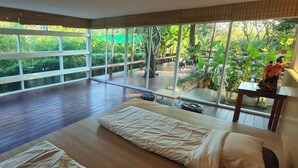 A spacious room with a large sliding glass door leading to a deck, wooden flooring, and a bed with a white comforter. - Natura Ravee Greenstay (Mae Taeng)