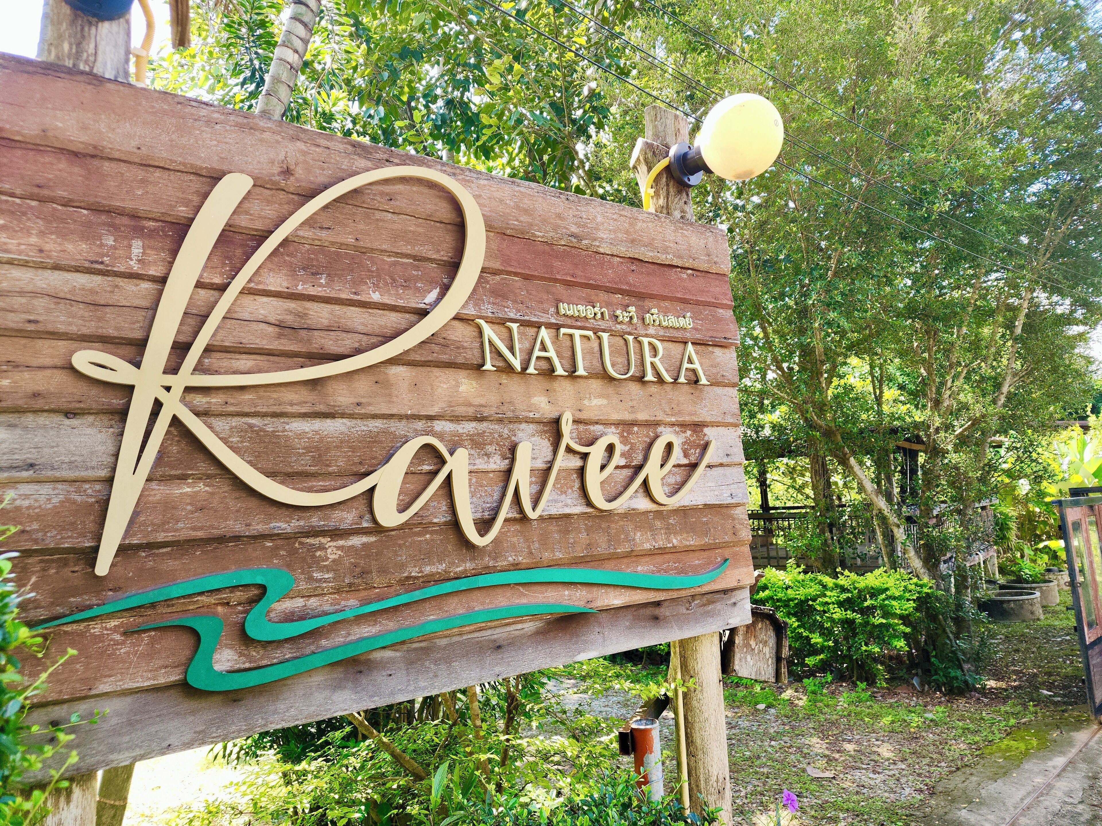 A wooden sign with the words "Natura" and "Ravee" in a stylized font, mounted on a post with a street lamp nearby.